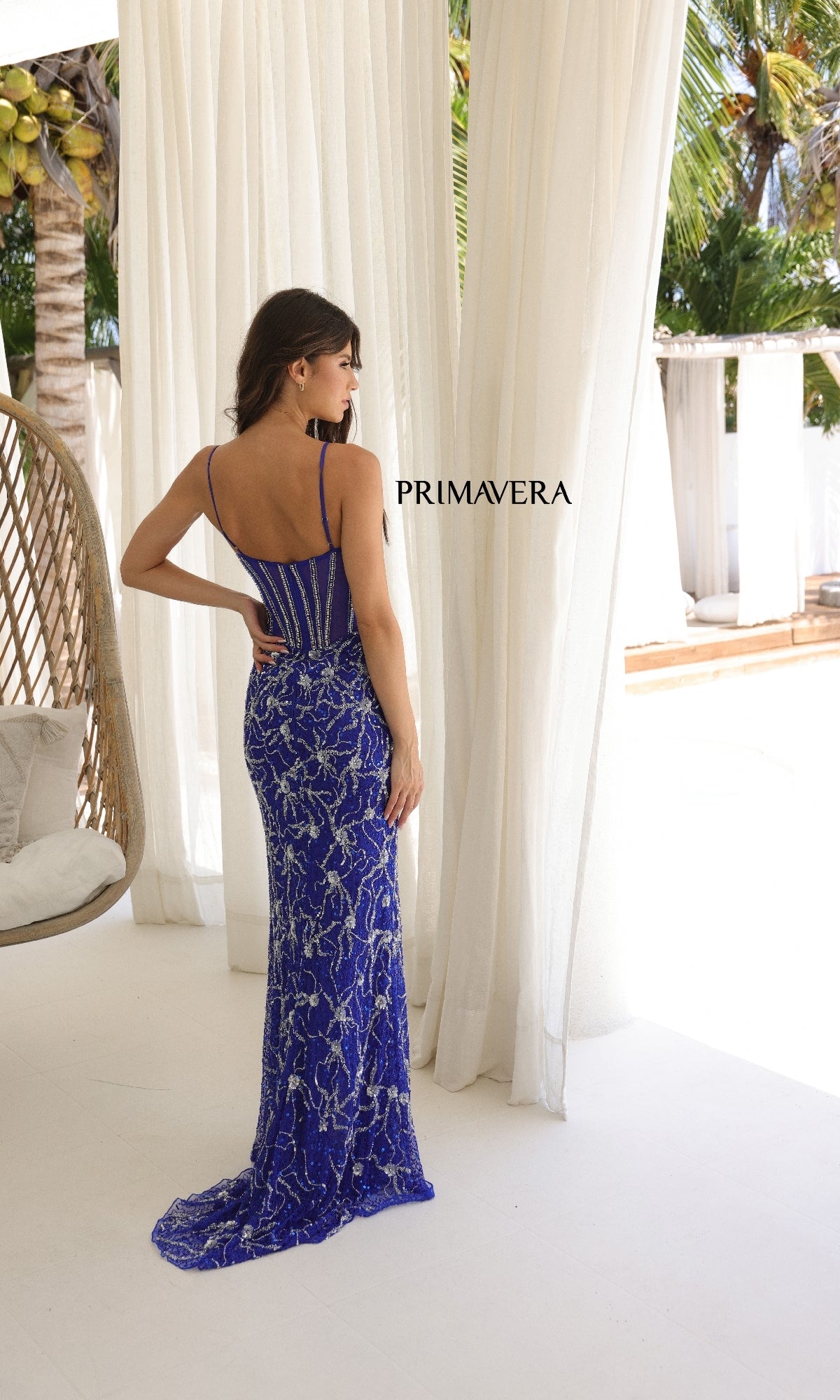 A woman in a fitted blue Sheer-Back Long Sequin Prom Dress (Primavera 4307) stands with her back to the camera in a sunny outdoor setting with white drapes and tropical plants. The word